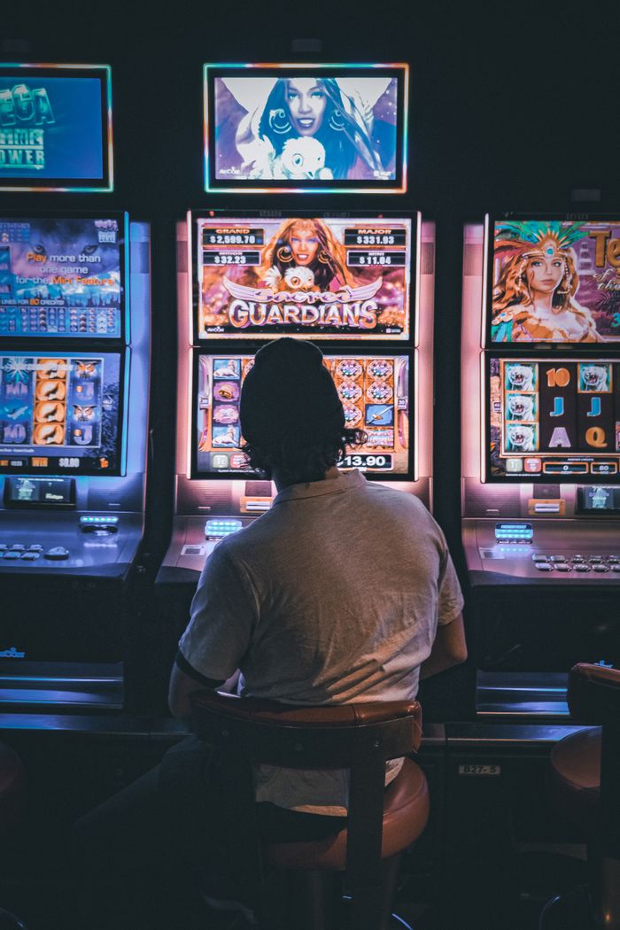 hero-img-01 Adult male seated in a dimly lit casino engaging with vibrant slot machines at night.